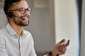 Um homem desempenhando a função de operador de telemarketing. Ele usa camisa social de cor clara, headset e óculos de grau.