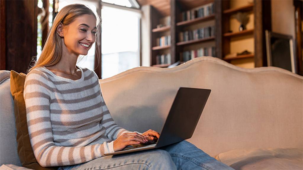 Uma mulher, em uma sala de estar, sentada no sofá e usando um notebook.