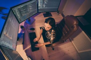 Uma mulher sentada à frente de uma mesa de madeira, com quatro telas de computadores, um teclado e um mouse.