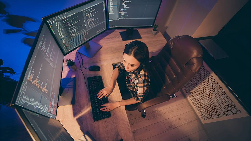 Uma mulher sentada à frente de uma mesa de madeira, com quatro telas de computadores, um teclado e um mouse.