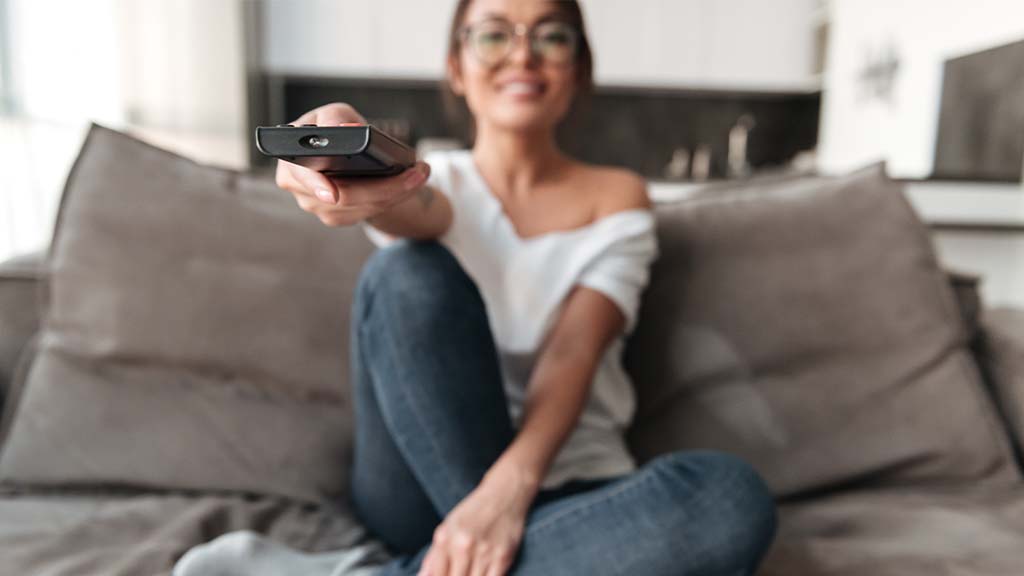 Uma mulher sentada no sofá segurando um controle remoto para ligar a TV.