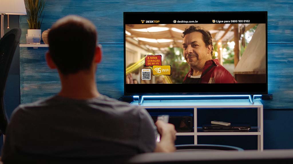 Um homem assistindo TV. Na tela do aparelho, uma cena do comercial da Desktop em que indica que a empresa oferece internet com o Wi-Fi 6.