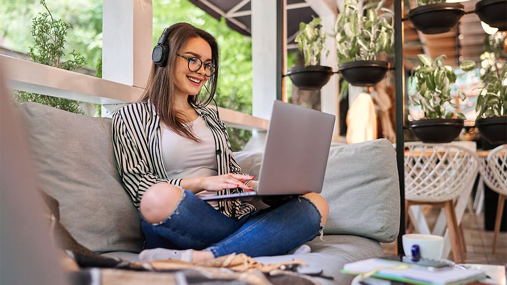 Uma mulher sentada no sofá, usando um notebook. Ela está de headphone. Ao lado dela vários vasos de plantas.