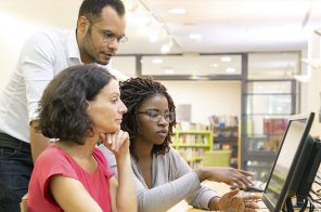 Três pessoas, sendo um homem em pé e duas mulheres sentadas, estão à frente de um computador conversando.