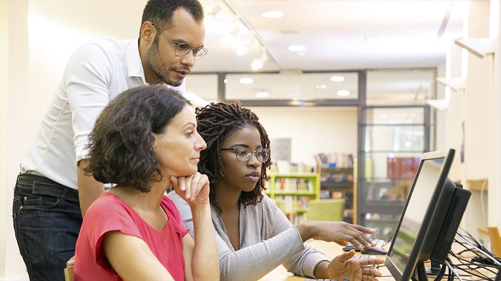 Três pessoas, sendo um homem em pé e duas mulheres sentadas, estão à frente de um computador conversando.